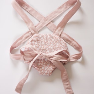 Peut inclure: Une barboteuse rose pour b&eacute;b&eacute; avec un motif floral blanc et des bretelles r&eacute;glables. Les bretelles sont nou&eacute;es en un n&oelig;ud au dos.