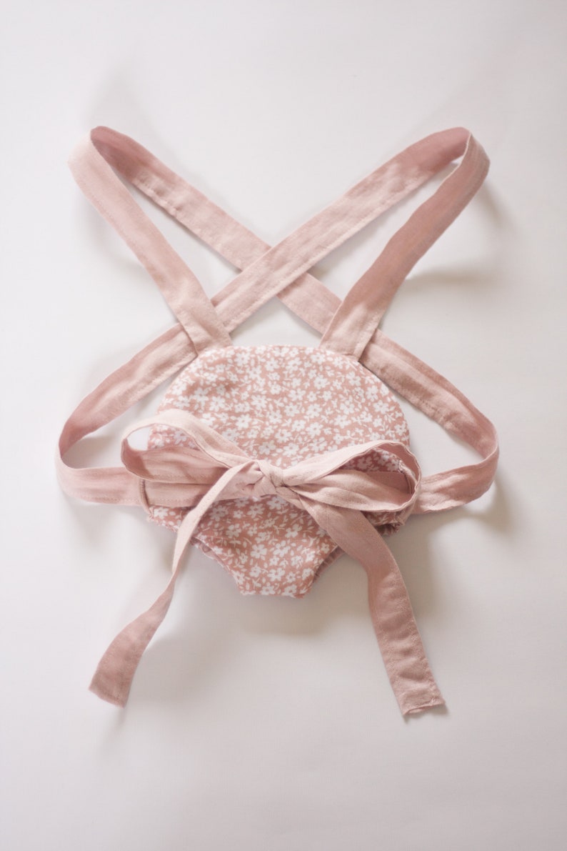 Peut inclure: Une barboteuse rose pour b&eacute;b&eacute; avec un motif floral blanc et des bretelles r&eacute;glables. Les bretelles sont nou&eacute;es en un n&oelig;ud au dos.