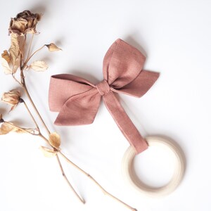 Peut inclure: Un jouet de dentition en forme de n&oelig;ud papillon rose poussi&eacute;reux avec un anneau en bois. Le n&oelig;ud est en tissu et est attach&eacute; &agrave; l'anneau par une bande de tissu. Le jouet est sur fond blanc avec des fleurs s&eacute;ch&eacute;es.