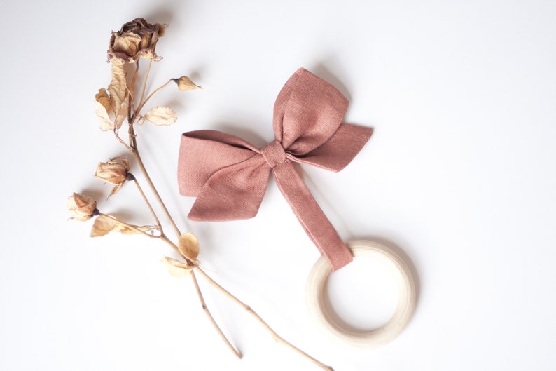 Peut inclure: Un jouet de dentition en forme de n&oelig;ud papillon rose poussi&eacute;reux avec un anneau en bois. Le n&oelig;ud est en tissu et est attach&eacute; &agrave; l'anneau par une bande de tissu. Le jouet est sur fond blanc avec des fleurs s&eacute;ch&eacute;es.