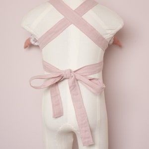 Peut inclure: Un porte-b&eacute;b&eacute; en lin rose avec des bretelles r&eacute;glables qui se croisent dans le dos et se nouent en un n&oelig;ud.