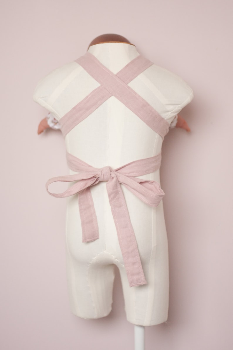Peut inclure: Un porte-b&eacute;b&eacute; en lin rose avec des bretelles r&eacute;glables qui se croisent dans le dos et se nouent en un n&oelig;ud.