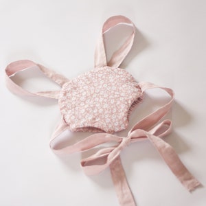 Peut inclure: Une barboteuse rose pour b&eacute;b&eacute; avec un motif floral blanc et des bretelles roses. La barboteuse a un n&oelig;ud papillon au dos.
