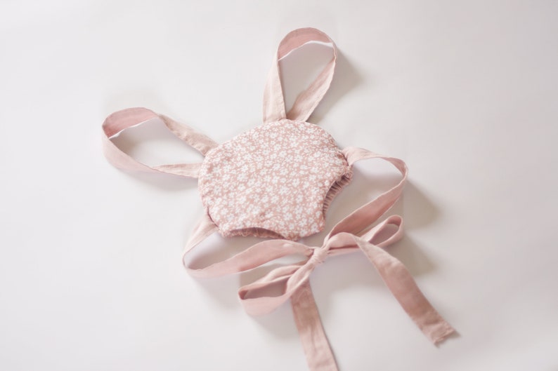 Peut inclure: Une barboteuse rose pour b&eacute;b&eacute; avec un motif floral blanc et des bretelles roses. La barboteuse a un n&oelig;ud papillon au dos.