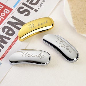 May include: Three curved, smooth, metallic objects with polished finishes. One is gold-toned with the name Robert engraved in script, while two are silver-toned with the names Dylan and Brooke engraved in script on their respective surfaces.