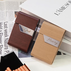 May include: Two rectangular cigarette cases with a wood-grain finish, one dark brown and one light tan. Each case features a diagonal silver-toned metal accent plate engraved with a name, specifically Jack on the dark case and Henry on the light case.