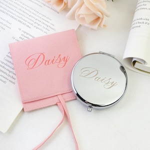 May include: A round silver-toned metal compact mirror and a square light pink suede-like pouch. Both items feature the name Daisy in a cursive script font. The mirror is polished and reflective, while the pouch includes a matching pink ribbon tie.
