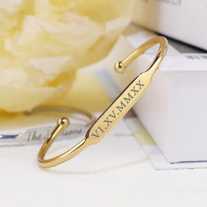 May include: A gold-colored cuff bracelet with rounded ends. The bracelet has the Roman numerals VI.XV.MMXX engraved on the front. The bracelet is a simple, elegant design.