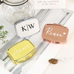 Puede incluir: Tres pastilleros rectangulares personalizados de metal en plata, oro rosa y oro. La caja plateada tiene "K/W" en negro. La caja de oro rosa tiene "Renee" con un corazón. La caja dorada tiene "Anne".
