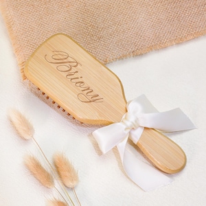 Può includere: Una spazzola per capelli rettangolare in legno chiaro, con il nome Briony inciso in corsivo sul retro. Il manico è decorato con un fiocco di nastro di raso bianco, che aggiunge un elemento decorativo alla venatura naturale del legno.