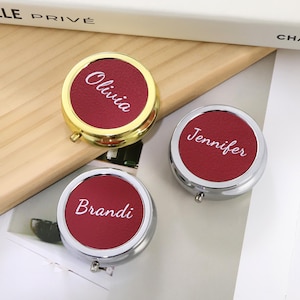 Op de afbeelding: Drie gepersonaliseerde pillendoosjes met namen in witte letters. Eén doosje heeft een gouden frame en de naam "Olivia". De andere twee hebben zilveren frames met de namen "Jennifer" en "Brandi" op een rode achtergrond.