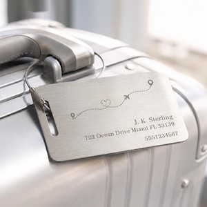 May include: A rectangular brushed metal luggage tag with a wire loop attachment. The tag features an engraved dashed flight path with a heart and airplane icon. It is personalized with the text J. K. Sterling, 722 Ocean Drive Miami FL 33139, 5551234567.
