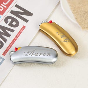 May include: Two curved metal lighters with a brushed finish, one in gold and one in silver. The gold lighter is engraved with the script text M.G. and the silver lighter is engraved with the script text Aaron. Both feature a red ignition button.