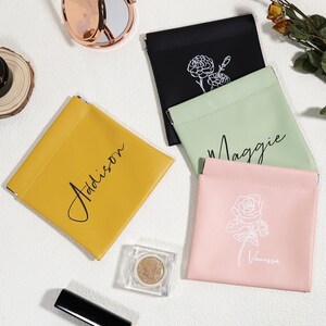 May include: An overhead shot displays four small, square pouches in yellow, black, light green, and pink. Each pouch has a name written on it in cursive. The black pouch has a floral design. A compact mirror and makeup are also visible.