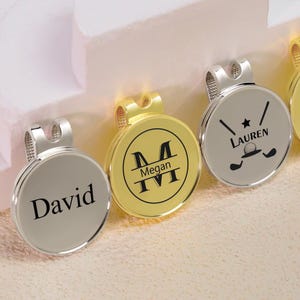 Könnte beinhalten: Nahaufnahme von drei personalisierten Golfballmarkern. Einer ist silberfarben mit dem Namen "David", ein anderer ist goldfarben mit "Megan IV", und der dritte ist silberfarben mit einem Golfmotiv und dem Namen "Lauren".