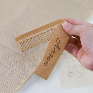 Könnte beinhalten: Ein hölzerner Klappkamm mit dem eingravierten Namen "Aaron" auf dem Griff. Der Kamm ist aus hellem Holz gefertigt und hat feine Zähne. Der Kamm ist geöffnet und zeigt die Zähne.