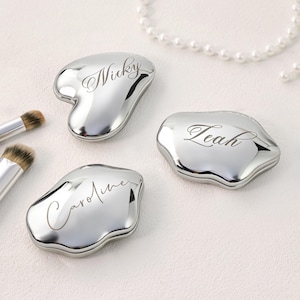 May include: Three silver-colored, personalized compact mirrors with engraved names: "Nicky", "Leah", and "Caroline". The mirrors have unique, organic shapes. Makeup brushes and a pearl necklace are also visible.