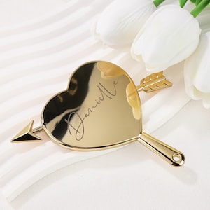 Pode incluir: Um espelho de mão dourado em forma de coração com um design de seta. O espelho tem o nome "Danielle" inscrito na superfície refletora. A pega tem um pequeno orifício. O espelho está colocado numa superfície branca com flores brancas no fundo.