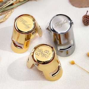 May include: Three metal champagne bottle stoppers with hinged locking mechanisms. Two are gold-toned and one is silver-toned. The gold stoppers are engraved with the name Hailey and the initials EB with a floral design and date 26. 8. 2026. The silver stopper is engraved with the name Taylor.