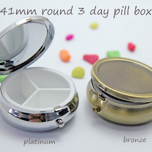 May include: Two 41mm round 3-day pill boxes, one platinum and one bronze. The platinum pill box is open, revealing a white interior divided into three sections. The bronze pill box is closed. Text on the image reads "platinum" and "bronze".