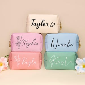 May include: A collection of personalized cosmetic bags in various pastel colors: cream, pink, light blue, and mint green. Each bag features a name in elegant script, with floral or heart accents. Gold zippers add a touch of detail.
