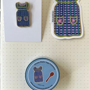 May include: A set of three items featuring a blue and purple gingham apron design. The set includes a sticker, a pin, and a roll of washi tape. The washi tape is 1.8cm x 10cm and features a spoon and apron design. The text on the washi tape reads "Abuelas Washi Tape Collection" and "www.abuelaskitchenofficial.com".