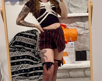 May include: A black crop top with a white crochet design, paired with red and black plaid shorts. The outfit is completed with fishnet stockings and black bows. The person is holding a phone in front of a mirror.