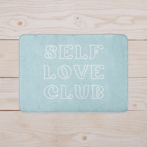 Pode incluir: Um tapete de banho azul claro com o texto "SELF LOVE CLUB" em letras brancas com contorno.