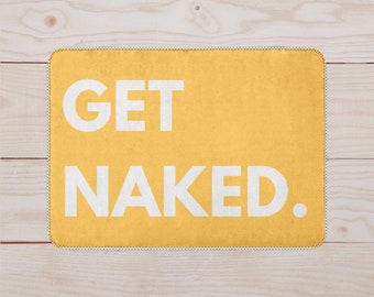 Op de afbeelding: Een gele badmat met de tekst "GET NAKED." in wit.