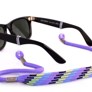Peut inclure: Lunettes de soleil noires avec un cordon de lunettes à motif violet et noir et une boucle en métal argenté.