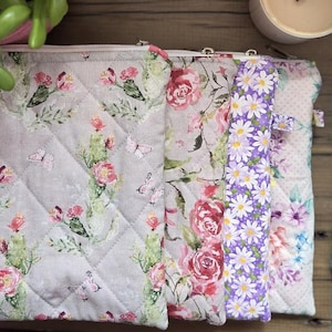May include: A collection of quilted fabric pouches with floral and butterfly patterns. The pouches feature a variety of designs, including roses, daisies, and other flowers in shades of pink, purple, and white. The pouches are arranged on a wooden surface.