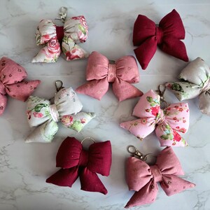 May include: A collection of fabric bow keychains in various colors and patterns. The bows are in shades of pink, red, and floral prints. Each bow has a metal clasp for attaching to keys or bags. The keychains are arranged on a white marble surface.
