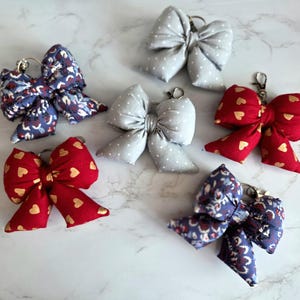 May include: A collection of handmade bow keychains in various colors and patterns. The bows are made of fabric and feature designs such as polka dots, floral prints, and heart patterns. Each keychain has a metal ring for attaching keys.