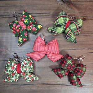 May include: Five fabric bow keychains in various patterns. The bows feature floral, plaid, and holly designs in green, red, and pink. Each bow has a silver-colored clasp for attaching to keys or bags.