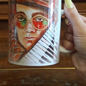 Może przedstawiać: Biały ceramiczny kubek z portretem osoby w czerwonych okularach i wzorem klawiatury fortepianu. Portret jest kolorowy z odcieniami czerwieni, pomarańczy i brązu. Kubek ma biały uchwyt i białe wnętrze.