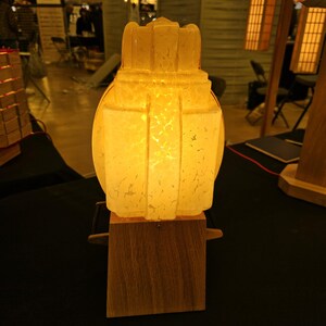 Peut inclure: Lampe décorative avec un abat-jour blanc cassé texturé et une base en bois. L'abat-jour présente un design architectural avec des panneaux verticaux et une forme arrondie. La lampe est éclairée, diffusant une lumière chaleureuse.