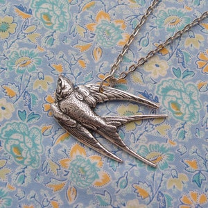 May include: A silver-colored pendant necklace featuring a detailed swallow in flight. The bird's wings are outstretched, and the chain is a delicate silver. The necklace is set against a light blue floral patterned background.