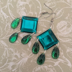 May include: A pair of dangle earrings featuring a large, square, teal-colored faceted crystal center piece. Each earring is accented with three smaller, teardrop-shaped, emerald green faceted crystals suspended from silver-toned metal jump rings.
