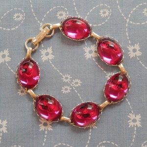 May include: A vintage bracelet with six oval, ruby-red glass stones set in a gold-tone metal frame. The stones are linked with gold-tone metal bars and a clasp. The bracelet is displayed on blue floral patterned fabric.