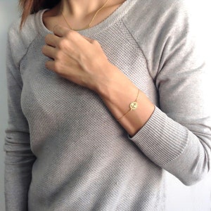 Puede incluir: Pulsera dorada con un dije circular con diseño de ancla. La pulsera se lleva en la muñeca. También se ve un collar dorado a juego. La persona lleva un suéter gris.