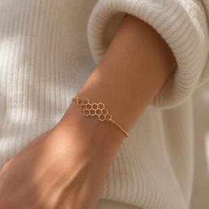 Puede incluir: Una pulsera de tono dorado que presenta un dije central con forma de panal compuesto por varios segmentos de alambre hexagonal interconectados. La delicada cadena de metal se conecta a cada lado de la pieza central geométrica de panal.