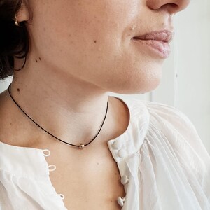 May include: A simple gold bead necklace on a black cord. The necklace is worn by a person with a white shirt.