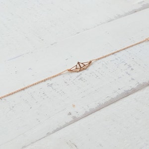 Puede incluir: Un brazalete de cadena dorada con un delicado dije de barco de origami. El brazalete está sobre una superficie de madera blanca.