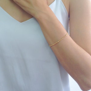 Puede incluir: Un delicado brazalete de cadena dorada con pequeñas cuentas redondas. El brazalete se lleva en la muñeca.