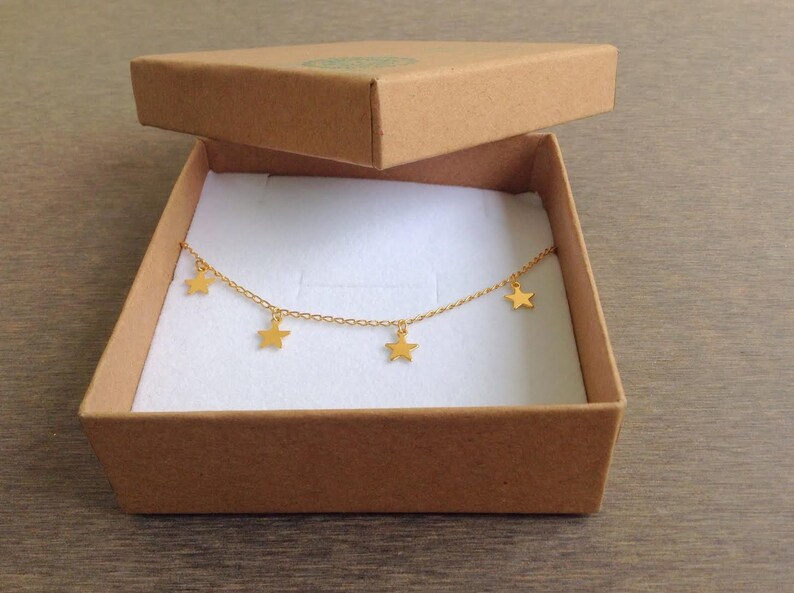 Puede incluir: Un collar de cadena dorada con cinco colgantes de estrellas en una caja de cart&oacute;n marr&oacute;n con interior blanco.