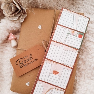 May include: A brown paper package with a book-themed bookmark. The bookmark has a pink background with a bookshelf design and a cup of coffee. The package has a tag that says "This Book Belongs To:"
