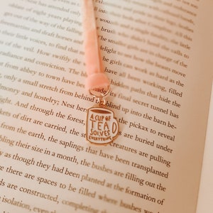 May include: A pink ribbon bookmark with a gold charm that says "A Cup of Tea Solves Everything". The bookmark is placed in an open book with text on the pages.