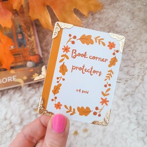 May include: A set of four gold book corner protectors with a fall-themed design. The protectors feature a white background with a wreath of orange and brown leaves, berries, and flowers. The text "Book corner protectors x4 gold" is printed in black.