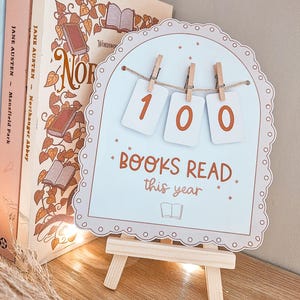 May include: A light blue sign on a wooden easel reads "100 BOOKS READ this year." The sign has a scalloped edge and is held by three small pegs. Books with floral designs are visible in the background.