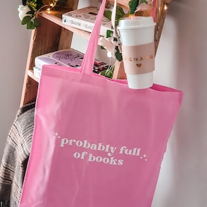 May include: Pink tote bag with the text "probably full of books" in white lettering. The bag is hanging on a wooden shelf with books and a coffee mug with a brown sleeve that says "mug in a mug" in gold lettering.